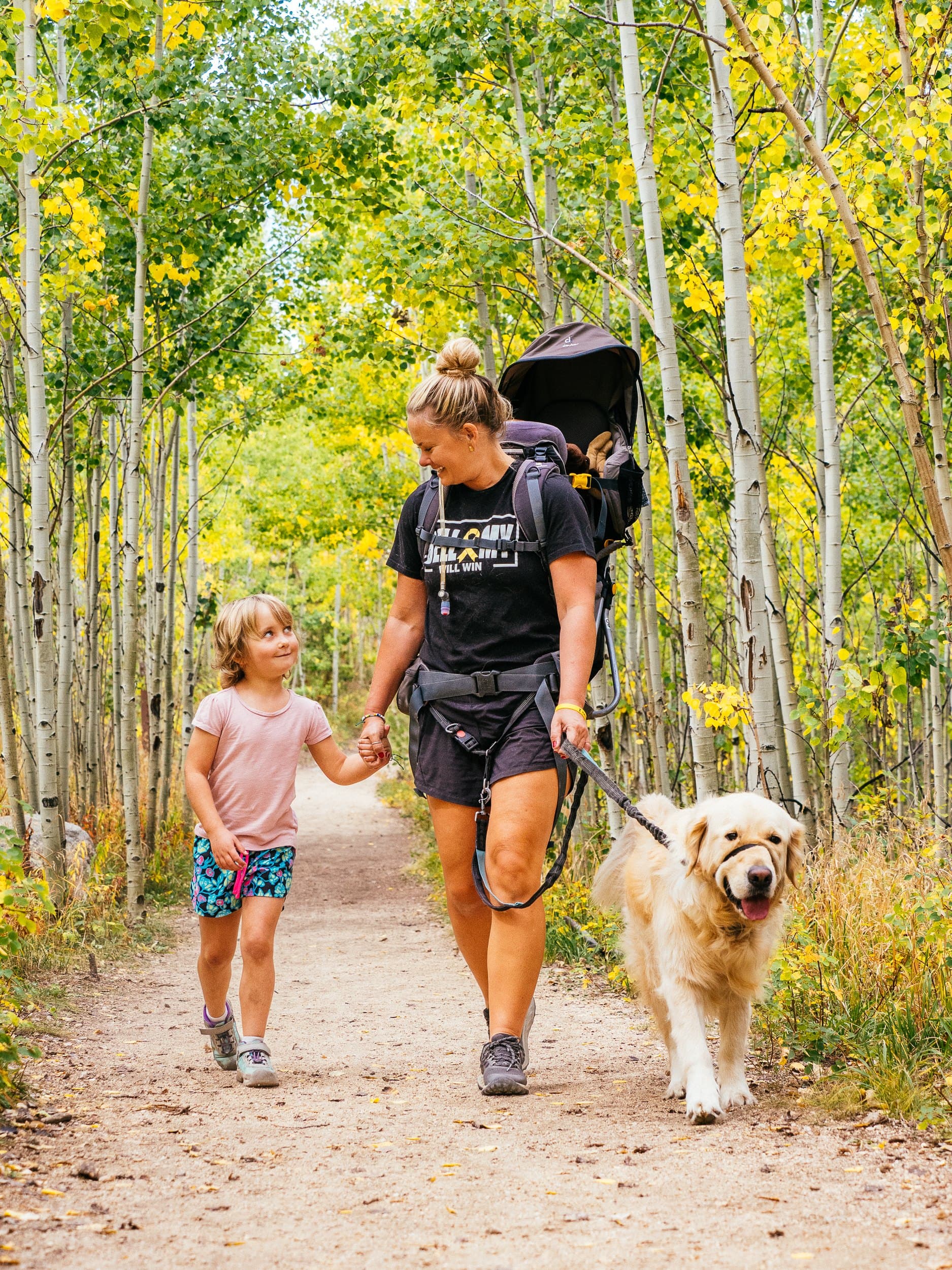 Sarah walking a golden aspen trail with her daughter, the baby in a carrier on her back and the dog alongside.