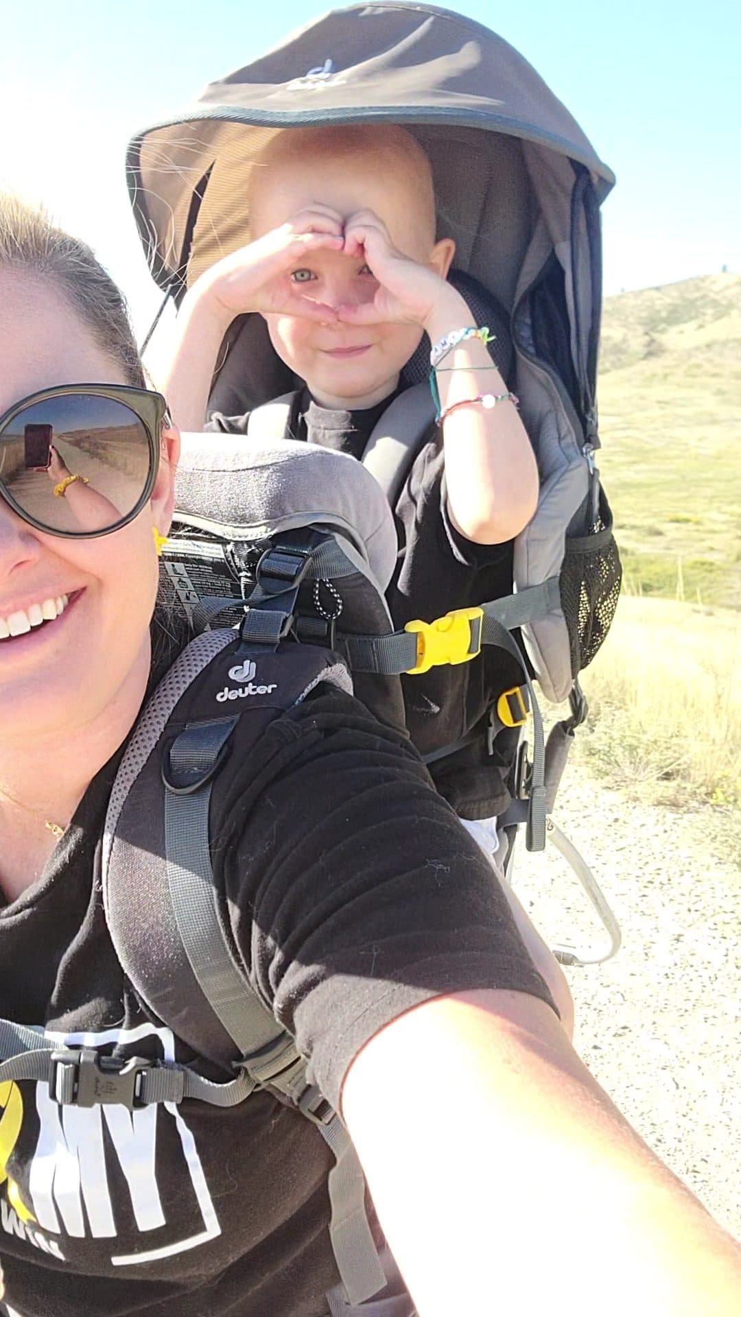Sarah smiling on the trail with Bellamy riding behind her in the old hiking backpack, hands framed over her eyes.
