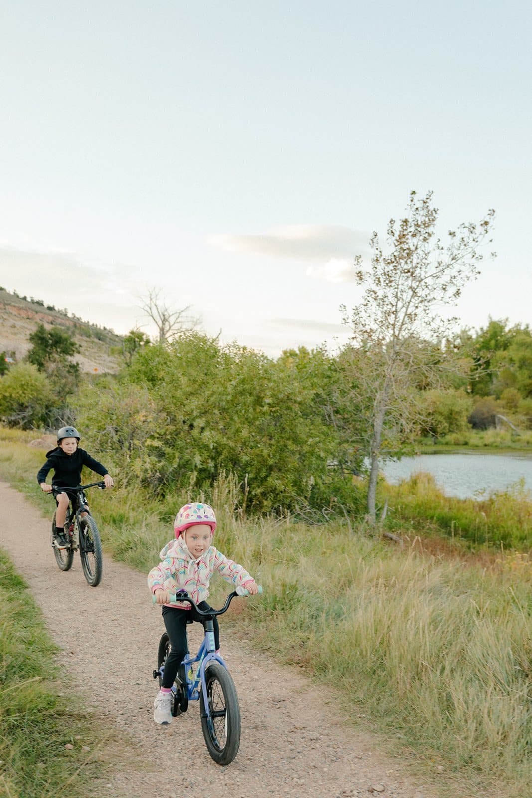 Two children riding bikes along a trail past a pond.