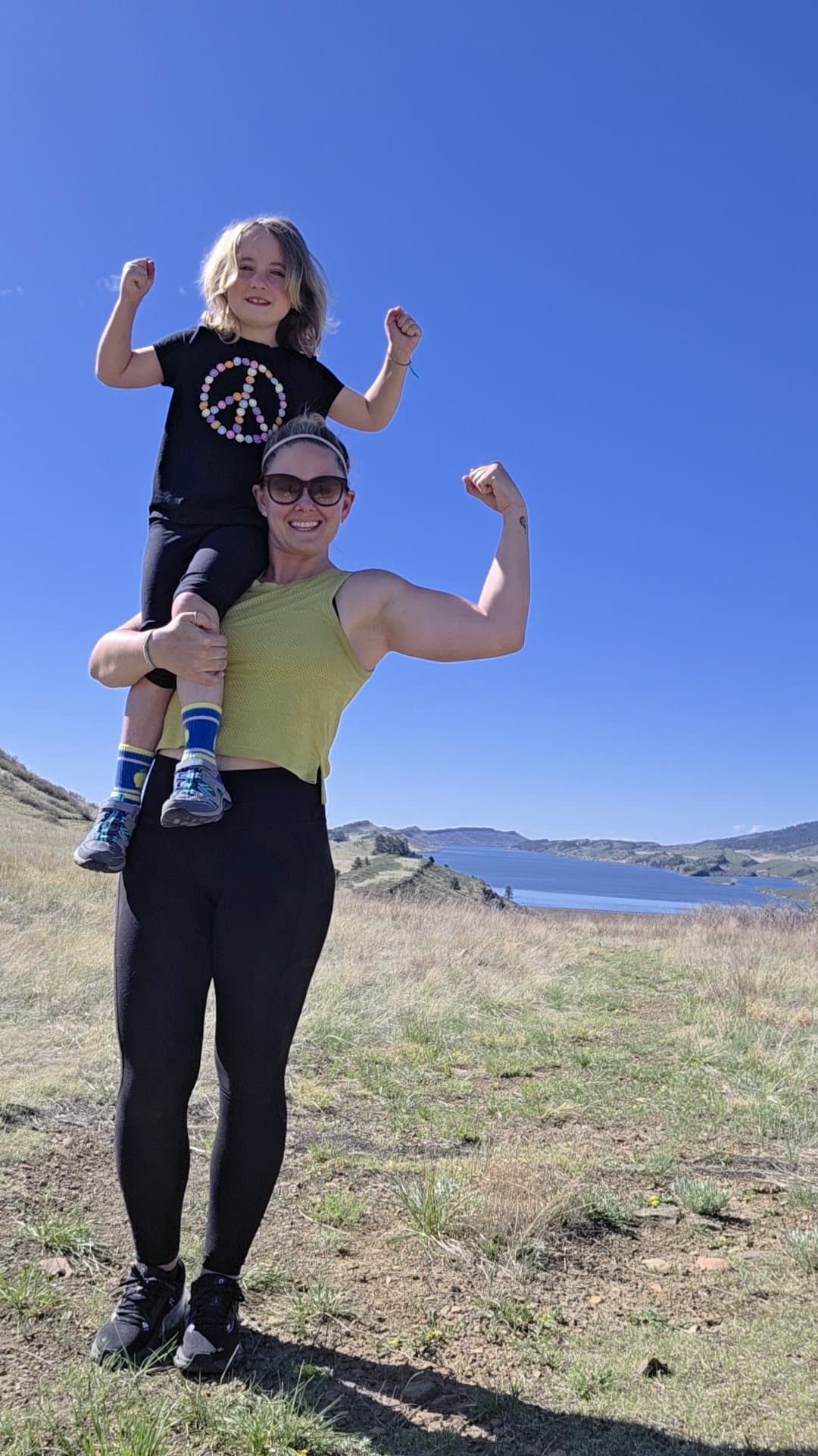Bellamy up on Sarah's shoulders, both of them flexing, under a blue sky.