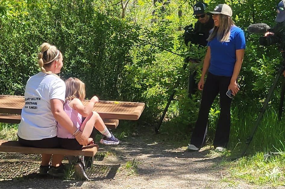 Sarah and Bellamy being interviewed on a bench at a trailhead, a reporter and camera in front of them.